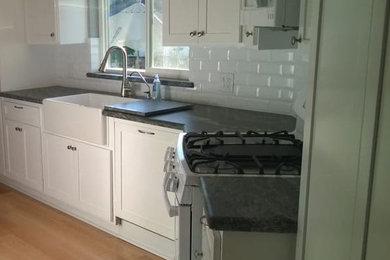 Mid-sized transitional l-shaped light wood floor and beige floor kitchen photo in Los Angeles with a farmhouse sink, recessed-panel cabinets, white cabinets, quartz countertops, white backsplash, ceramic backsplash, white appliances, no island and black countertops