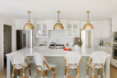 Inspiration for a large transitional l-shaped eat-in kitchen in Boston with a farmhouse sink, recessed-panel cabinets, white cabinets, quartz benchtops, white splashback, ceramic splashback, stainless steel appliances, light hardwood floors, with island and white benchtop.