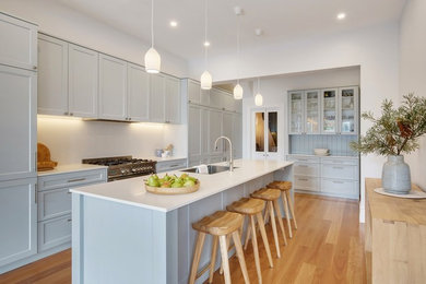 Mid-sized beach style kitchen in Sydney with a single-bowl sink, shaker cabinets, grey cabinets, solid surface benchtops, white splashback, stainless steel appliances, with island, white benchtop, light hardwood floors and beige floor.