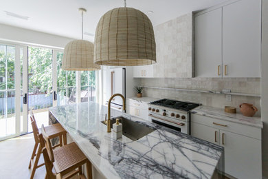 Mid-sized trendy single-wall light wood floor and brown floor open concept kitchen photo in New York with a drop-in sink, flat-panel cabinets, white cabinets, marble countertops, beige backsplash, ceramic backsplash, white appliances, an island and multicolored countertops