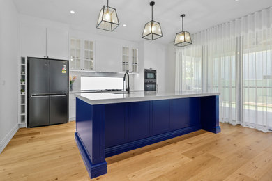 This is an example of a large galley open plan kitchen in Sydney with an undermount sink, shaker cabinets, blue cabinets, quartz benchtops, white splashback, engineered quartz splashback, black appliances, laminate floors, with island, brown floor and white benchtop.