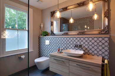 Photo of a mid-sized contemporary bathroom in Kent with flat-panel cabinets, light wood cabinets, an open shower, a wall-mount toilet, gray tile, porcelain tile, grey walls, porcelain floors, a vessel sink and laminate benchtops.