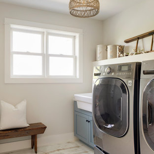 ミネアポリスの中サイズのモダンスタイルのおしゃれな洗濯室 (エプロンフロントシンク、インセット扉のキャビネット、青いキャビネット、ベージュの壁、塗装フローリング、左右配置の洗濯機・乾燥機、白い床) の写真