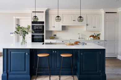 Large traditional open plan kitchen in Buckinghamshire with an integrated sink, raised-panel cabinets, blue cabinets, quartz worktops, white splashback, black appliances, medium hardwood flooring, an island and white worktops.