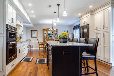 Large transitional u-shaped medium tone wood floor, brown floor and vaulted ceiling kitchen pantry photo in Boston with an island, raised-panel cabinets, white cabinets, granite countertops, gray countertops, gray backsplash, subway tile backsplash, stainless steel appliances and an undermount sink