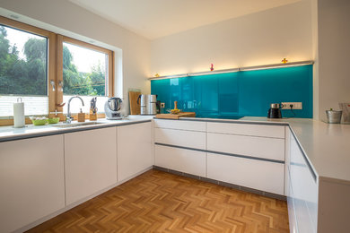 Inspiration for a mid-sized contemporary u-shaped separate kitchen in Cologne with medium hardwood floors, white cabinets, an integrated sink, flat-panel cabinets, wood benchtops, blue splashback, glass sheet splashback, no island and brown floor.