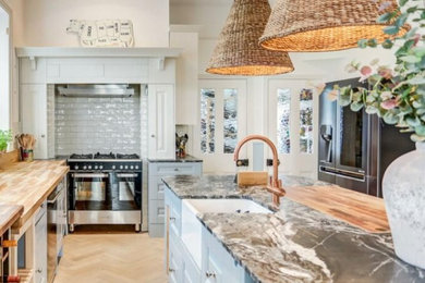This is an example of a large bohemian l-shaped kitchen/diner in Sussex with a belfast sink, beaded cabinets, blue cabinets, marble worktops, stainless steel appliances, light hardwood flooring, an island, grey worktops and feature lighting.