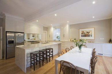 This is an example of an expansive contemporary l-shaped eat-in kitchen in Sydney with recessed-panel cabinets, white cabinets, mirror splashback, stainless steel appliances, light hardwood floors and with island.
