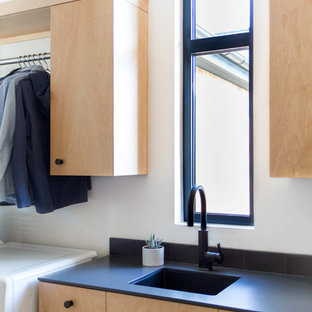 Réalisation d'une grande buanderie parallèle minimaliste multi-usage avec un évier encastré, un placard à porte plane, des portes de placard en bois clair, un plan de travail en surface solide, un mur blanc, béton au sol et des machines côte à côte.