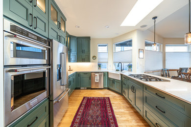 Mid-sized transitional u-shaped medium tone wood floor and brown floor open concept kitchen photo in Seattle with a farmhouse sink, green cabinets, white backsplash, stainless steel appliances, white countertops, flat-panel cabinets, quartz countertops, ceramic backsplash and a peninsula