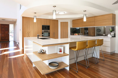 This is an example of a large modern l-shaped open plan kitchen in Brisbane with flat-panel cabinets, light wood cabinets, mirror splashback, stainless steel appliances, with island, brown floor, an undermount sink, quartz benchtops and medium hardwood floors.