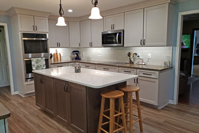Example of a mid-sized transitional l-shaped vinyl floor and brown floor eat-in kitchen design in Nashville with a farmhouse sink, recessed-panel cabinets, gray cabinets, quartz countertops, gray backsplash, ceramic backsplash, stainless steel appliances, an island and brown countertops