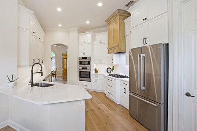 Example of a large transitional galley medium tone wood floor and brown floor kitchen design in Jacksonville with shaker cabinets, white cabinets, quartz countertops, white backsplash, porcelain backsplash, a peninsula, white countertops, an undermount sink and stainless steel appliances