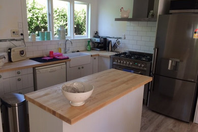 Traditional separate kitchen in Christchurch with a farmhouse sink, flat-panel cabinets, white cabinets, wood benchtops, white splashback, ceramic splashback, stainless steel appliances, light hardwood floors and with island.