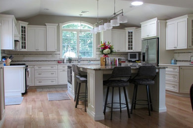 Large transitional u-shaped light wood floor and brown floor kitchen photo in New York with an undermount sink, recessed-panel cabinets, white cabinets, solid surface countertops, white backsplash, subway tile backsplash, stainless steel appliances and an island