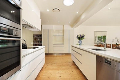 Design ideas for a mid-sized traditional galley eat-in kitchen in Newcastle - Maitland with a drop-in sink, flat-panel cabinets, white cabinets, quartz benchtops, beige splashback, glass sheet splashback, black appliances, light hardwood floors, with island and white benchtop.