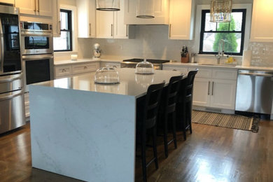 Example of a mid-sized trendy l-shaped medium tone wood floor eat-in kitchen design in Boston with an undermount sink, shaker cabinets, white cabinets, quartz countertops, white backsplash, porcelain backsplash, stainless steel appliances, an island and white countertops