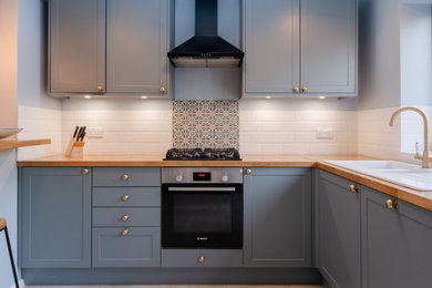 Inspiration for a medium sized traditional l-shaped open plan kitchen in London with a built-in sink, recessed-panel cabinets, blue cabinets, wood worktops, white splashback, ceramic splashback, black appliances, ceramic flooring, a breakfast bar, grey floors and a feature wall.
