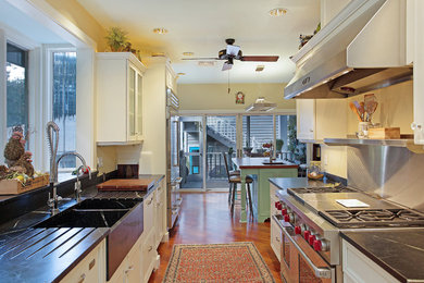 Mid-sized elegant galley medium tone wood floor eat-in kitchen photo in Tampa with a farmhouse sink, recessed-panel cabinets, white cabinets, soapstone countertops, beige backsplash, stone slab backsplash, stainless steel appliances and an island
