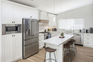 Example of a transitional l-shaped vaulted ceiling open concept kitchen design in Vancouver with an undermount sink, shaker cabinets, white cabinets, white backsplash, stainless steel appliances, an island and white countertops