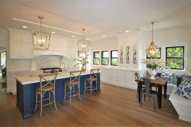 This is an example of a traditional l-shaped eat-in kitchen in Los Angeles with white cabinets, shaker cabinets, marble benchtops, white splashback, stainless steel appliances, light hardwood floors, with island and a farmhouse sink.