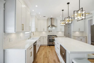 This is an example of a large classic u-shaped open plan kitchen in New York with a belfast sink, shaker cabinets, white cabinets, composite countertops, white splashback, marble splashback, stainless steel appliances, medium hardwood flooring, an island, brown floors and white worktops.