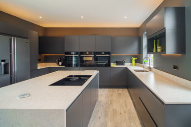Medium sized modern grey and black u-shaped kitchen/diner in Glasgow with a built-in sink, flat-panel cabinets, grey cabinets, granite worktops, grey splashback, granite splashback, black appliances, light hardwood flooring, an island, grey floors and grey worktops.