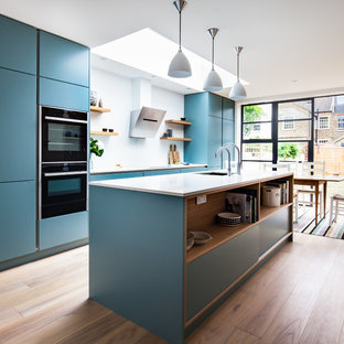 Ejemplo de cocina comedor lineal, actual, grande, con fregadero bajoencimera, armarios con paneles lisos, puertas de armario azules, encimera de cuarcita, salpicadero blanco, salpicadero de vidrio templado, electrodomésticos negros, suelo de madera en tonos medios y una isla