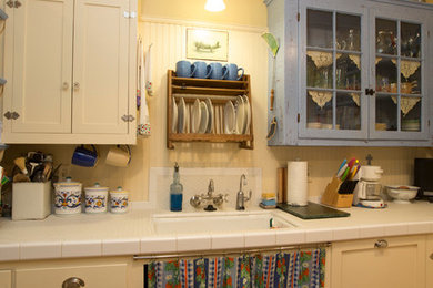 Inspiration for a small shabby-chic style l-shaped light wood floor and brown floor enclosed kitchen remodel in Other with an undermount sink, shaker cabinets, white cabinets, tile countertops, white backsplash, wood backsplash, white appliances and an island