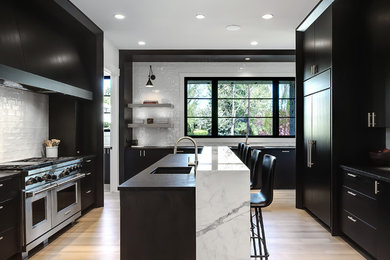 This is an example of a mid-sized contemporary l-shaped open plan kitchen in Calgary with an undermount sink, flat-panel cabinets, black cabinets, marble benchtops, white splashback, ceramic splashback, black appliances, medium hardwood floors, multiple islands and brown floor.