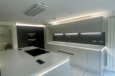 Inspiration for a large modern grey and white open plan kitchen in Oxfordshire with a built-in sink, flat-panel cabinets, white cabinets, grey splashback, glass sheet splashback, integrated appliances, ceramic flooring, an island, white floors, white worktops and feature lighting.