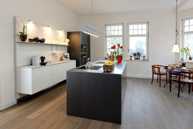 Large contemporary galley eat-in kitchen in Bremen with an undermount sink, flat-panel cabinets, black cabinets, stainless steel benchtops, white splashback, timber splashback, black appliances, dark hardwood floors, with island and brown floor.