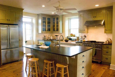 Example of a country l-shaped light wood floor open concept kitchen design in Other with a farmhouse sink, raised-panel cabinets, green cabinets, granite countertops, white backsplash, ceramic backsplash, stainless steel appliances and an island