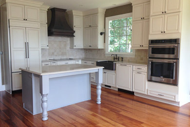 Example of a transitional l-shaped medium tone wood floor eat-in kitchen design in Charleston with a farmhouse sink, raised-panel cabinets, distressed cabinets, solid surface countertops, beige backsplash, stone tile backsplash, stainless steel appliances and an island