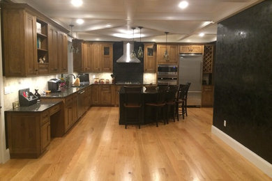 Example of a large classic l-shaped light wood floor eat-in kitchen design in Boston with a double-bowl sink, shaker cabinets, medium tone wood cabinets, granite countertops, black backsplash, ceramic backsplash, stainless steel appliances and an island