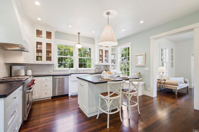 Mid-sized country l-shaped medium tone wood floor eat-in kitchen photo in San Francisco with a farmhouse sink, shaker cabinets, white cabinets, granite countertops, white backsplash, ceramic backsplash, stainless steel appliances, an island and black countertops