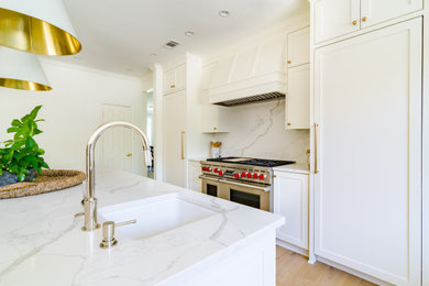 This is an example of a medium sized traditional kitchen in Dallas with a belfast sink, recessed-panel cabinets, white cabinets, engineered stone countertops, white splashback, engineered quartz splashback, stainless steel appliances, light hardwood flooring, an island, brown floors and white worktops.