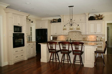 Example of a mid-sized classic medium tone wood floor and beige floor eat-in kitchen design in Other with a farmhouse sink, raised-panel cabinets, white cabinets, granite countertops, beige backsplash, porcelain backsplash, black appliances and an island