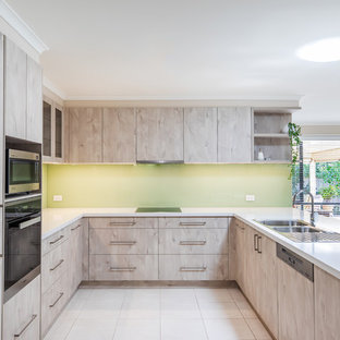 Inspiration for a mid-sized modern u-shaped kitchen pantry in Adelaide with a double-bowl sink, flat-panel cabinets, light wood cabinets, quartz benchtops, green splashback, glass sheet splashback, stainless steel appliances, porcelain floors, a peninsula, white floor and white benchtop.