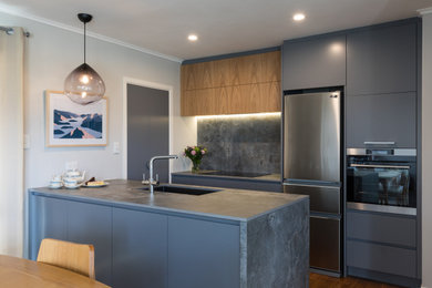 Photo of a small contemporary galley eat-in kitchen in Auckland with a double-bowl sink, flat-panel cabinets, grey cabinets, grey splashback, porcelain splashback, black appliances, light hardwood floors, a peninsula, brown floor and grey benchtop.