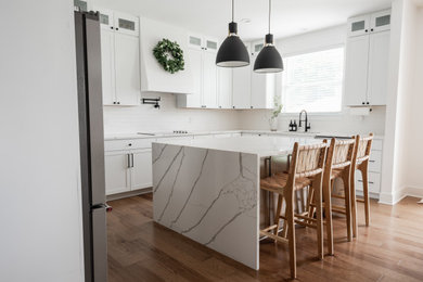 Mid-sized trendy l-shaped medium tone wood floor eat-in kitchen photo in Detroit with a single-bowl sink, shaker cabinets, white cabinets, quartz countertops, white backsplash, ceramic backsplash, stainless steel appliances, an island and white countertops