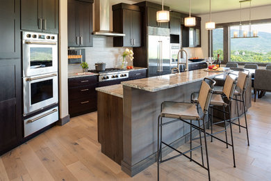 This is an example of a mid-sized modern u-shaped eat-in kitchen in Denver with a farmhouse sink, shaker cabinets, dark wood cabinets, quartzite benchtops, glass tile splashback, stainless steel appliances, light hardwood floors, multiple islands, brown floor, grey splashback and multi-coloured benchtop.