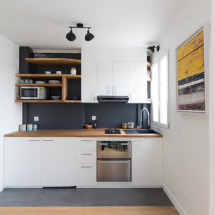 Пример оригинального дизайна интерьера: маленькая линейная кухня в стиле модернизм с обеденным столом, одинарной раковиной, белыми фасадами, столешницей из дерева, черным фартуком, фартуком из сланца, техникой под мебельный фасад, черным полом, коричневой столешницей, плоскими фасадами и бетонным полом без острова