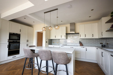 Photo of a large modern l-shaped kitchen/diner in Kent with a single-bowl sink, shaker cabinets, white cabinets, quartz worktops, blue splashback, ceramic splashback, integrated appliances, an island, brown floors and white worktops.