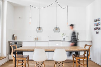Einzeilige, Große Moderne Wohnküche ohne Insel mit flächenbündigen Schrankfronten, weißen Schränken, Mineralwerkstoff-Arbeitsplatte, Küchenrückwand in Weiß, Rückwand aus Porzellanfliesen, Küchengeräten aus Edelstahl, braunem Holzboden, braunem Boden, weißer Arbeitsplatte und Unterbauwaschbecken in Berlin