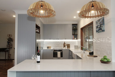Photo of a large country u-shaped separate kitchen in Brisbane with a farmhouse sink, flat-panel cabinets, blue cabinets, quartz benchtops, white splashback, subway tile splashback, stainless steel appliances, medium hardwood floors, a peninsula, brown floor and white benchtop.