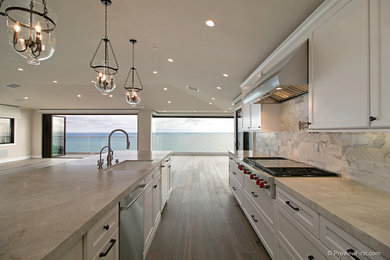 Design ideas for a large beach style l-shaped separate kitchen in Orange County with an undermount sink, shaker cabinets, white cabinets, white splashback, stone tile splashback, stainless steel appliances, medium hardwood floors, with island, quartzite benchtops and white benchtop.