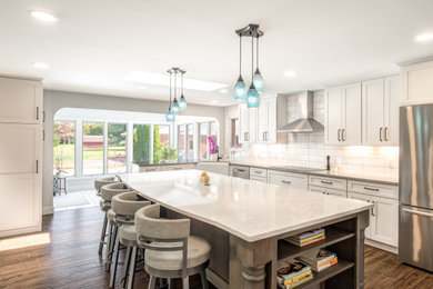 This is an example of a large transitional l-shaped open plan kitchen in Other with a farmhouse sink, shaker cabinets, white cabinets, quartz benchtops, white splashback, ceramic splashback, stainless steel appliances, with island, brown floor, white benchtop and porcelain floors.