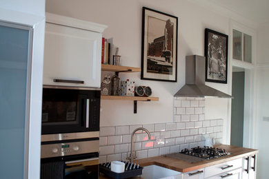Large rustic galley kitchen/diner in Glasgow with a belfast sink, shaker cabinets, white cabinets, wood worktops, white splashback, ceramic splashback, black appliances, ceramic flooring and a breakfast bar.