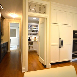 Cette photo montre une grande arrière-cuisine chic en U avec un évier encastré, un placard avec porte à panneau encastré, des portes de placard blanches, un plan de travail en verre, une crédence beige, une crédence en carreau de ciment, un électroménager encastrable, un sol en bois brun et 2 îlots.